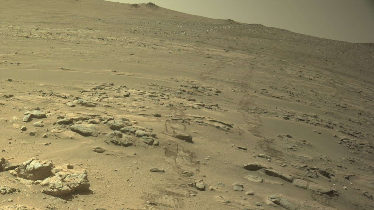 Rover wheel tracks in the sand of a rocky Martian slope leading toward a distant ridge under a hazy sky, seen by Perseverance's navigation camera on October 11, 2026